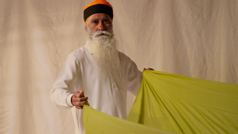 Fotografía-De-Estudio-De-Un-Hombre-Sikh-Mayor-Doblando-Tela-Para-Un-Turbante-Contra-Un-Fondo-Liso-Como-Secuencia,-Parte-1-De-2